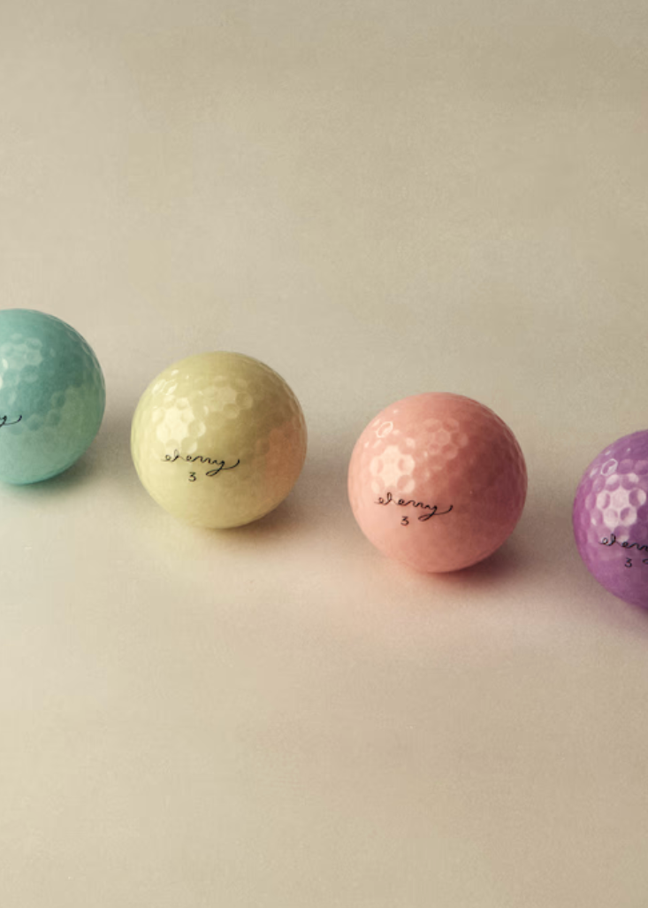 Four mini golf balls in pastel colors—mint green, pale yellow, light pink, and purple—arranged in a row on a warm beige background. Each ball has the brand name 'shiny' printed on it in black script.