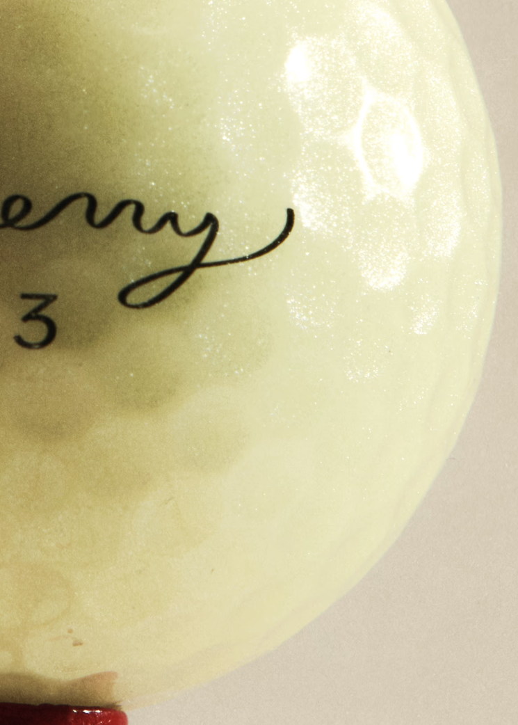 Close-up of a golf ball with partial black script lettering and the number 3, photographed against a plain warm beige background.