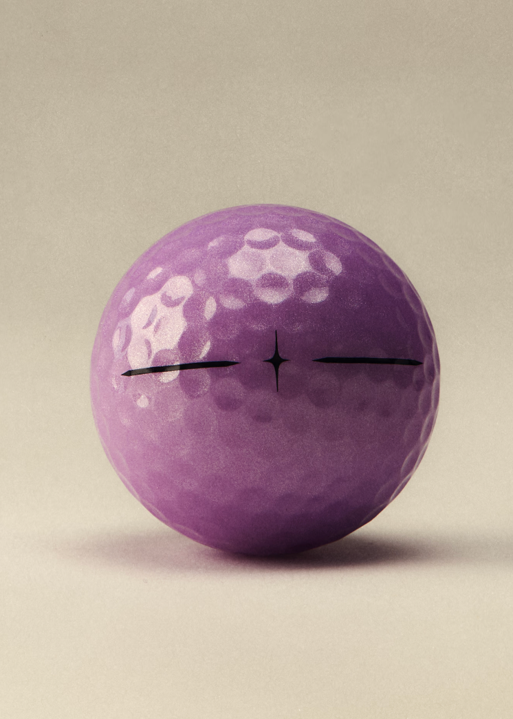 A single purple golf ball with black crosshair markings on a warm beige background.