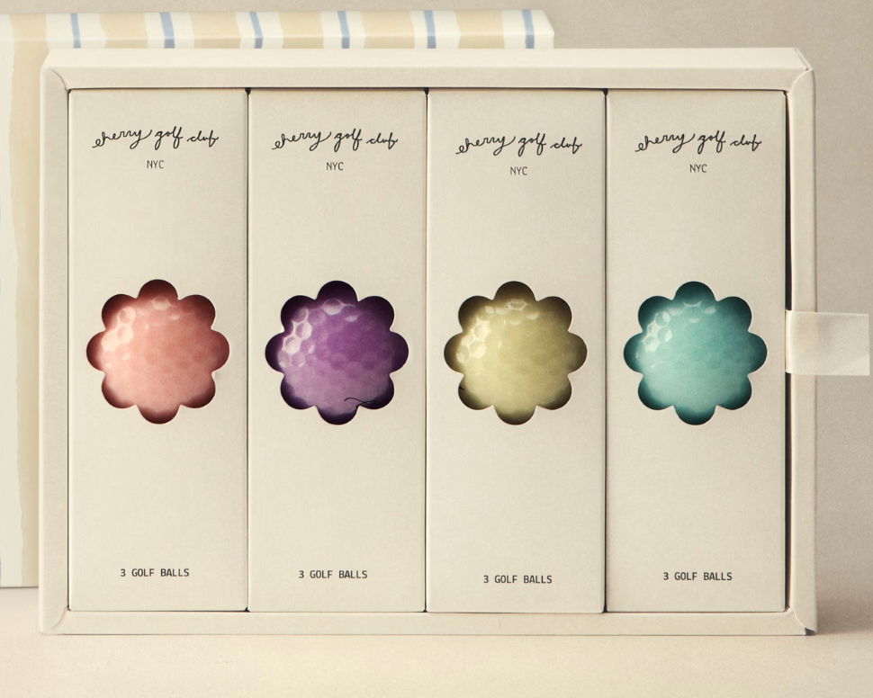 A boxed set of four golf ball packages arranged in a white rectangular box, each package featuring a flower-shaped cutout showing the golf ball color: pink, purple, yellow, and blue. The packages are labeled 'gravy golf clubs NYC' and '3 GOLF BALLS'.