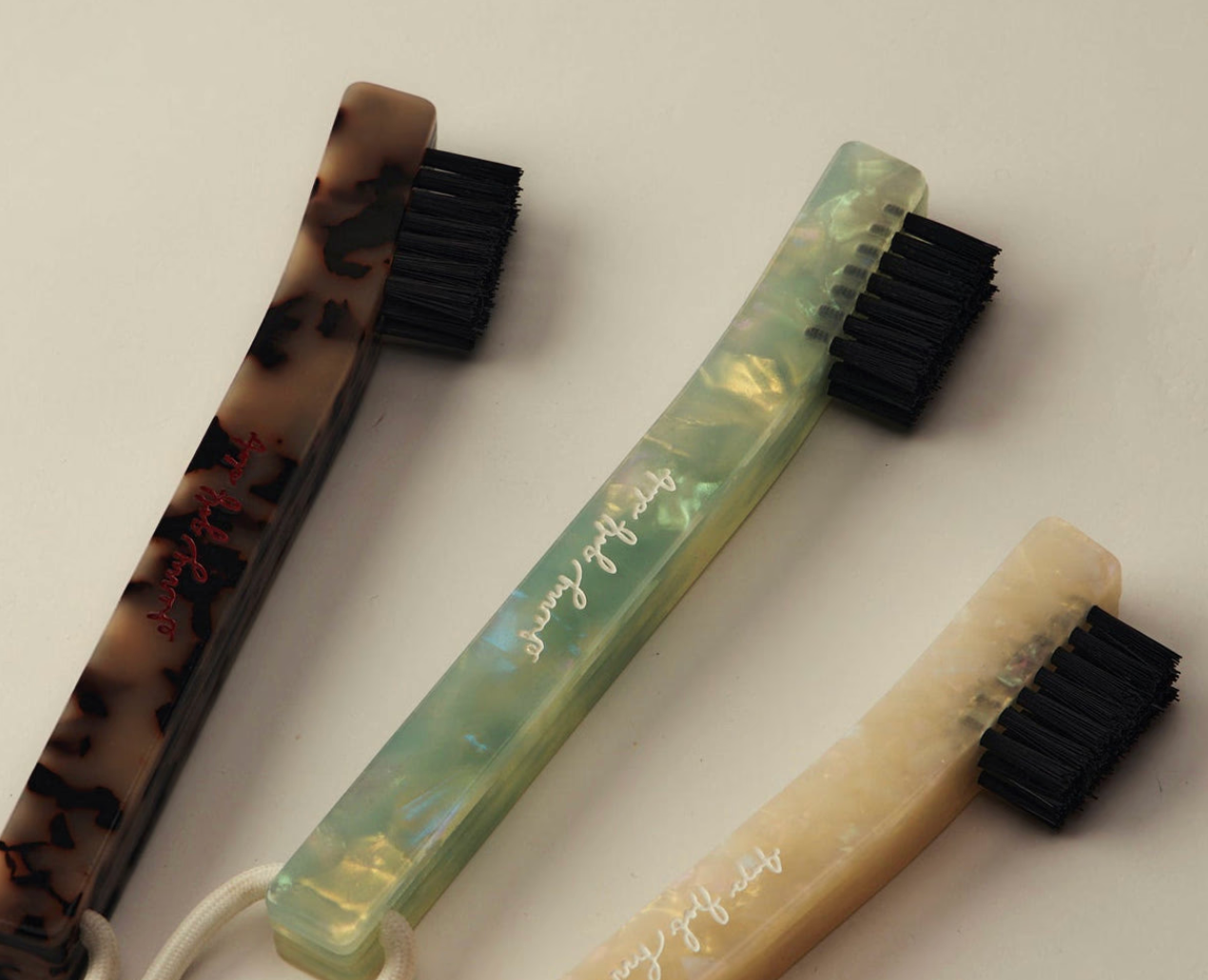 Three toothbrushes with decorative handles in tortoiseshell, iridescent green, and cream, arranged diagonally on a light beige background.