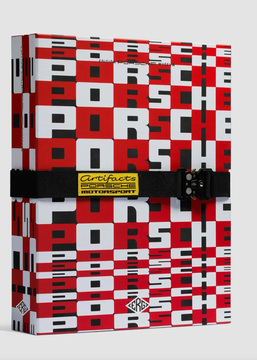 A Porsche-branded hardcover notebook or journal featuring a bold red, white, and black checkerboard pattern. It is secured with a black elastic band across the middle, which has a yellow and black Certificate of Authenticity label.