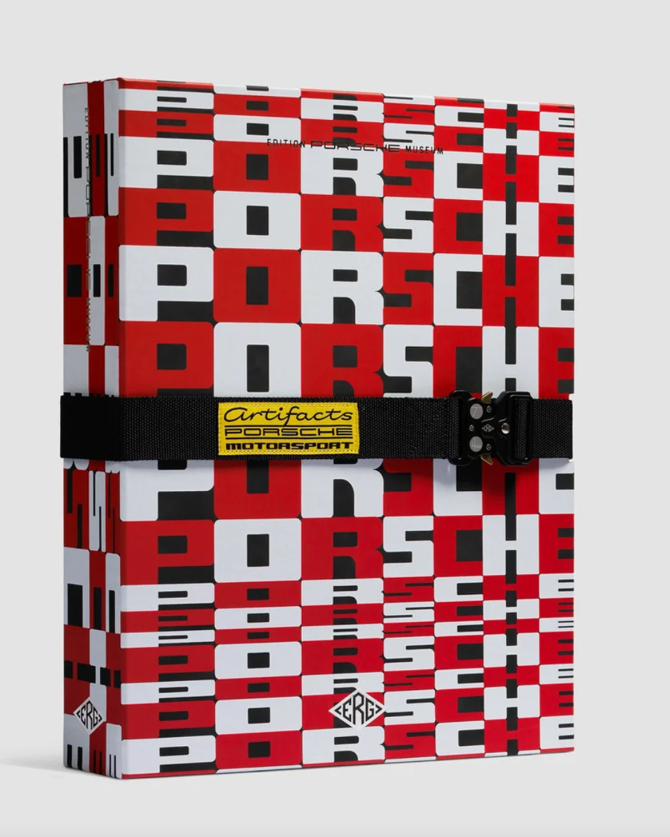 A Porsche-branded hardcover notebook or journal featuring a bold red, white, and black checkerboard pattern. It is secured with a black elastic band across the middle, which has a yellow and black Certificate of Authenticity label.