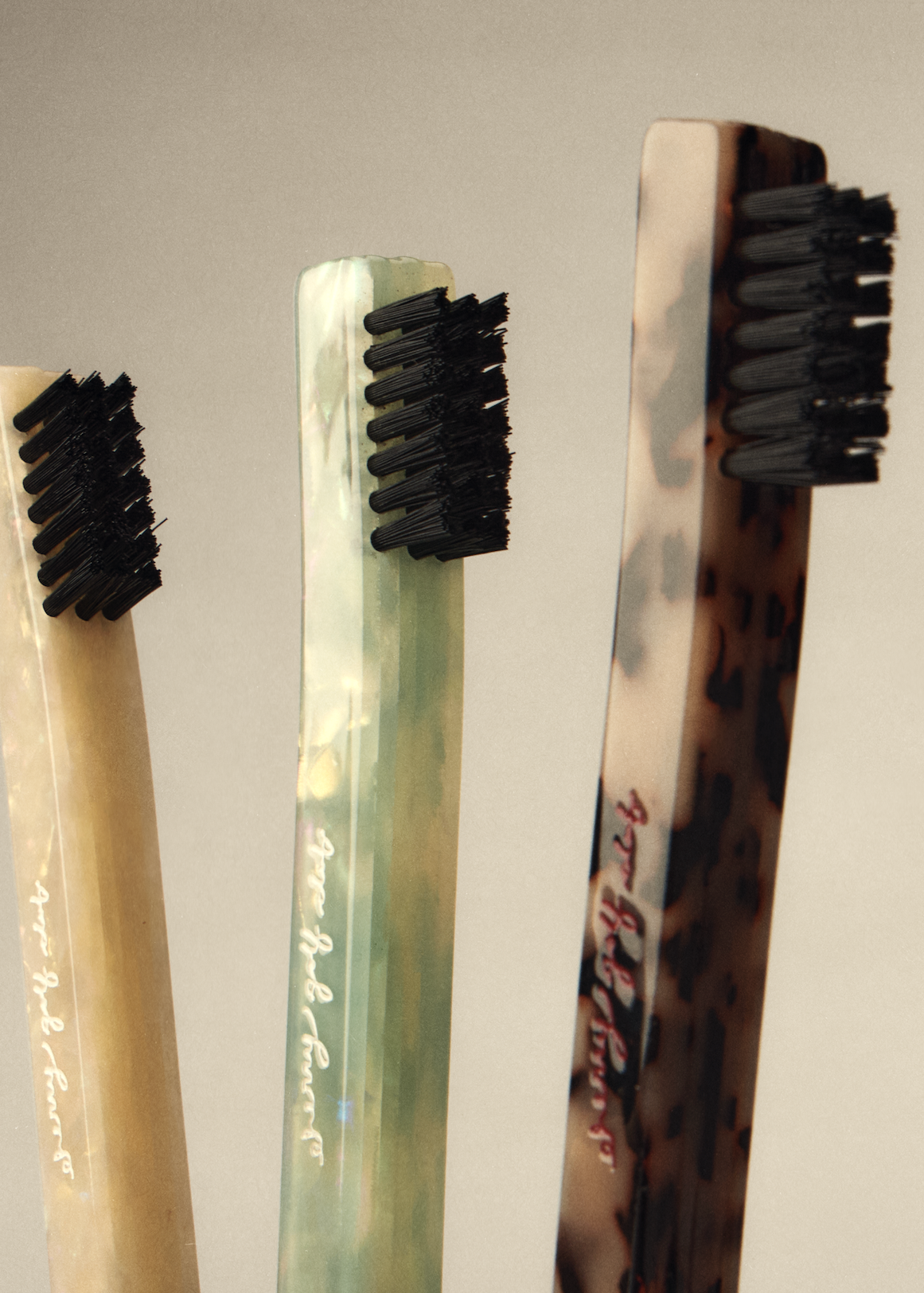 Three decorative toothbrushes with black bristles and marbled handles in beige, green, and brown, standing upright against a neutral gray background.