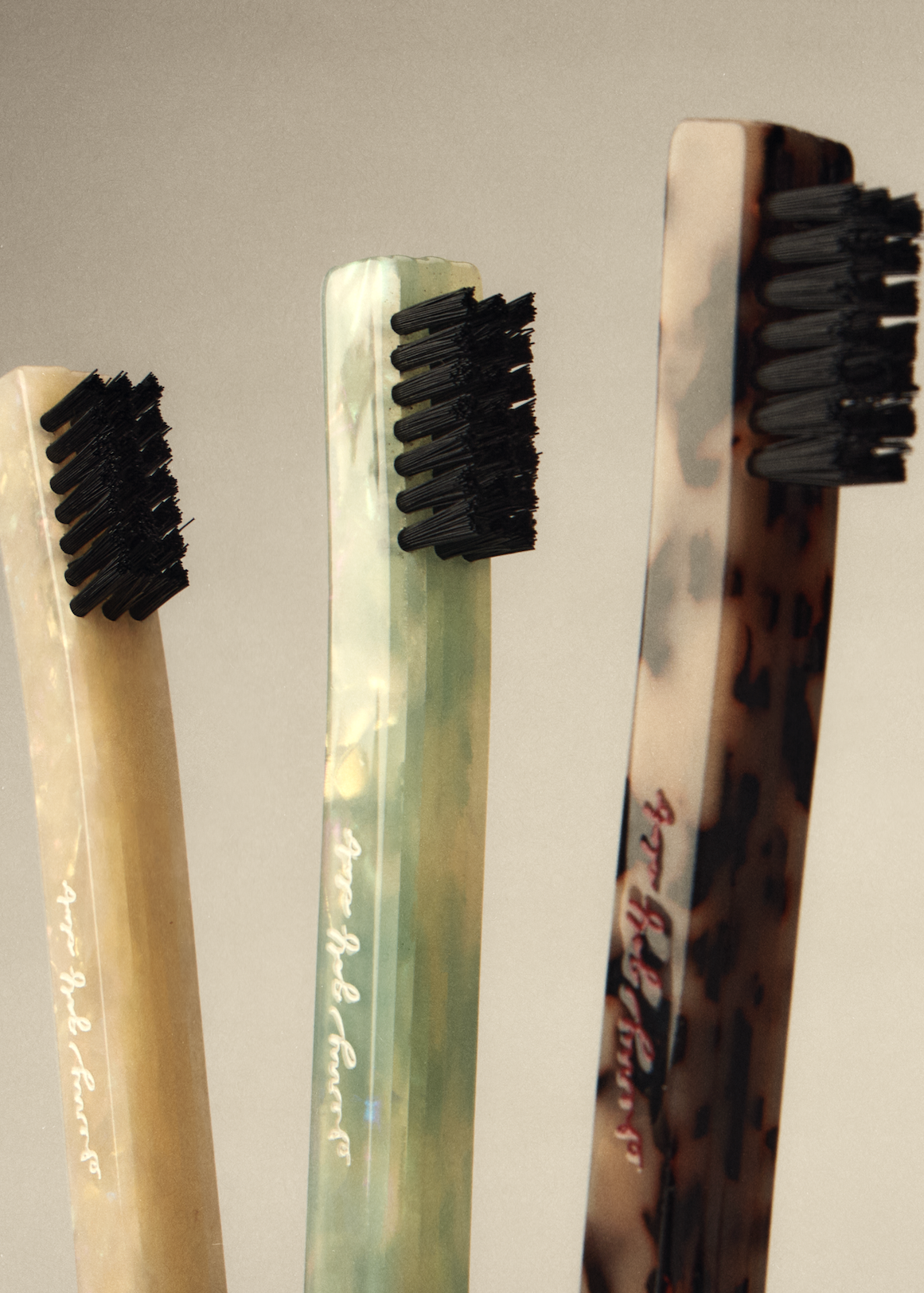 Three decorative toothbrushes with black bristles and marbled handles in beige, green, and brown, standing upright against a neutral gray background.