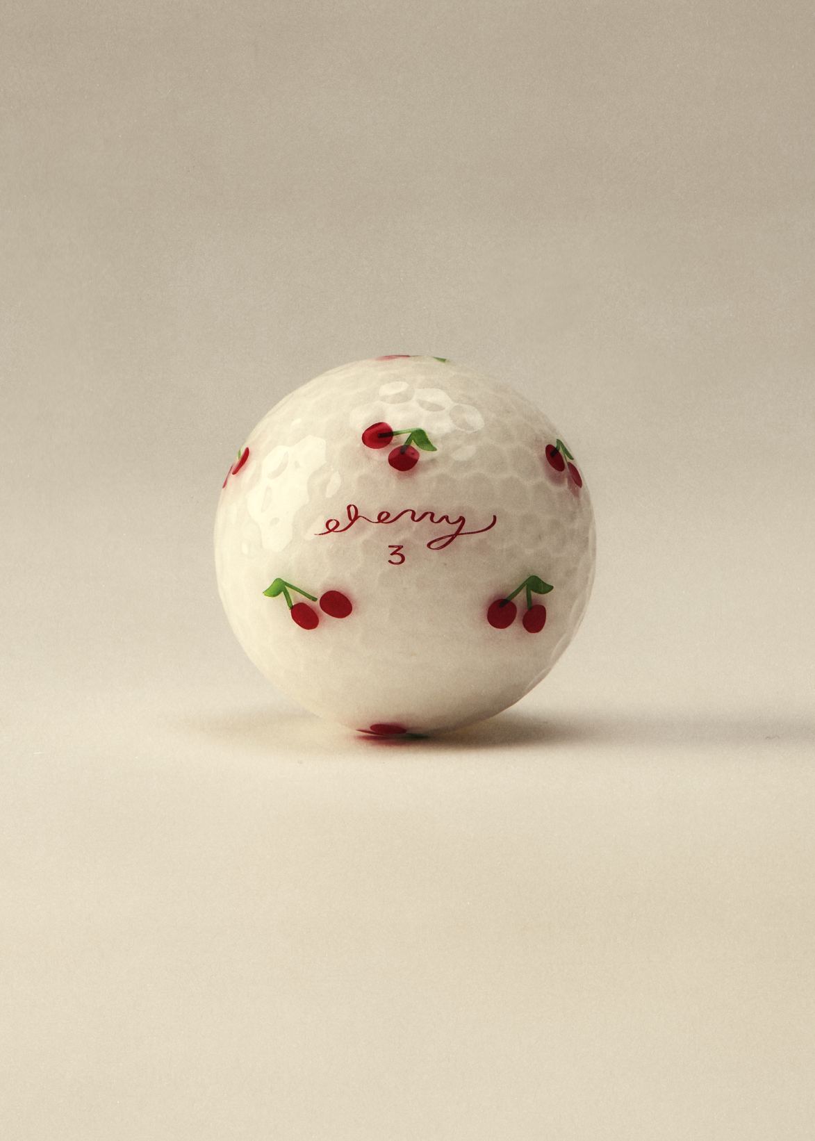 A white golf ball with a hand-painted cherry pattern and the cursive brand name 'cherry' printed on the front, set against a solid light beige background.