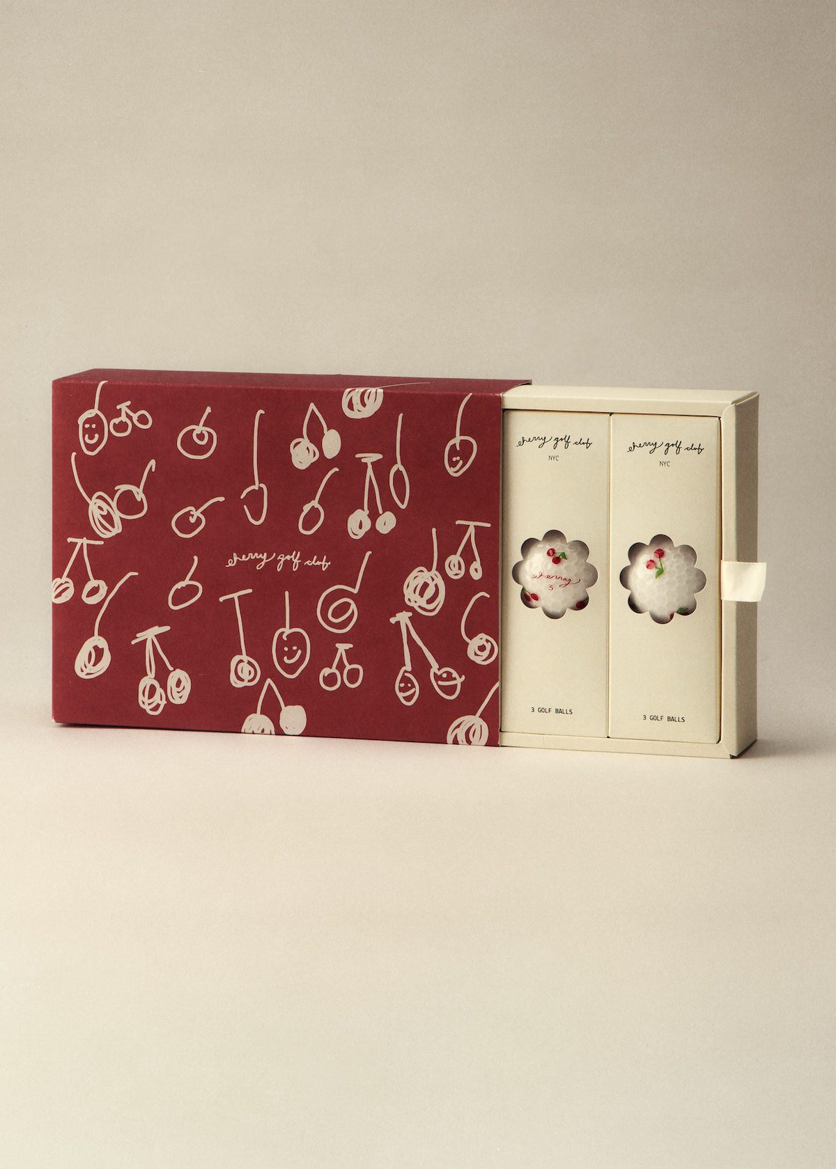 A boxed set of two small round pastries in cream-colored packaging, shown next to a larger dark red outer box decorated with white cherry illustrations, on a plain warm beige studio background.