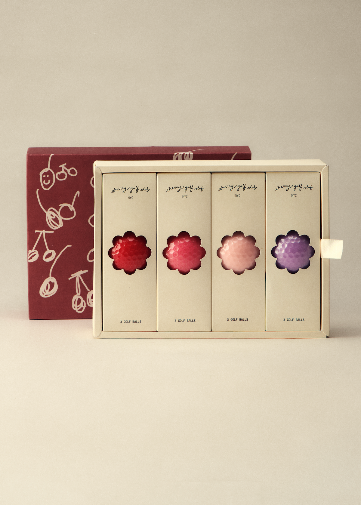 A boxed set of four flower-shaped cosmetic or fragrance items displayed in a cream-colored tray, with a dark red patterned box behind it on a light beige studio background.