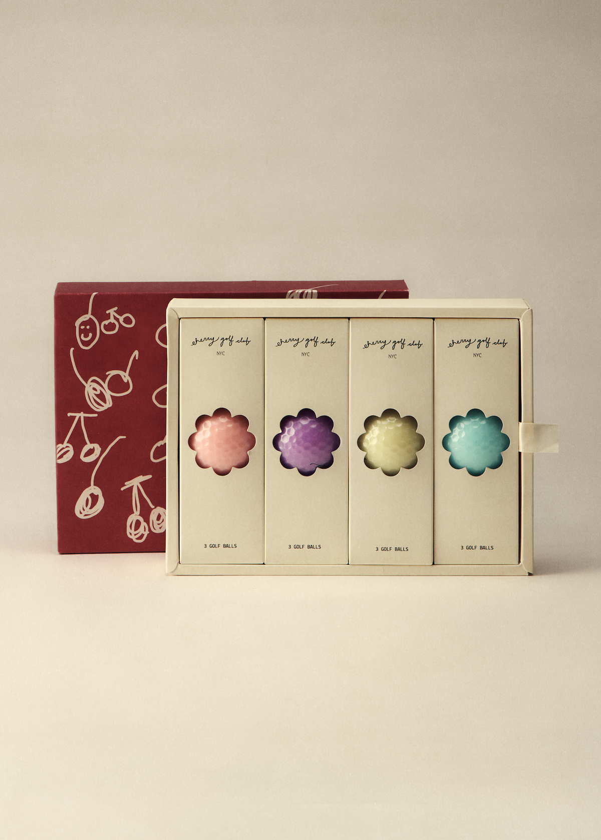 A boxed set of four flower-shaped cosmetic or candy pieces in pastel pink, purple, yellow, and blue, displayed in front of a dark red box with white cherry illustrations on a warm beige background.