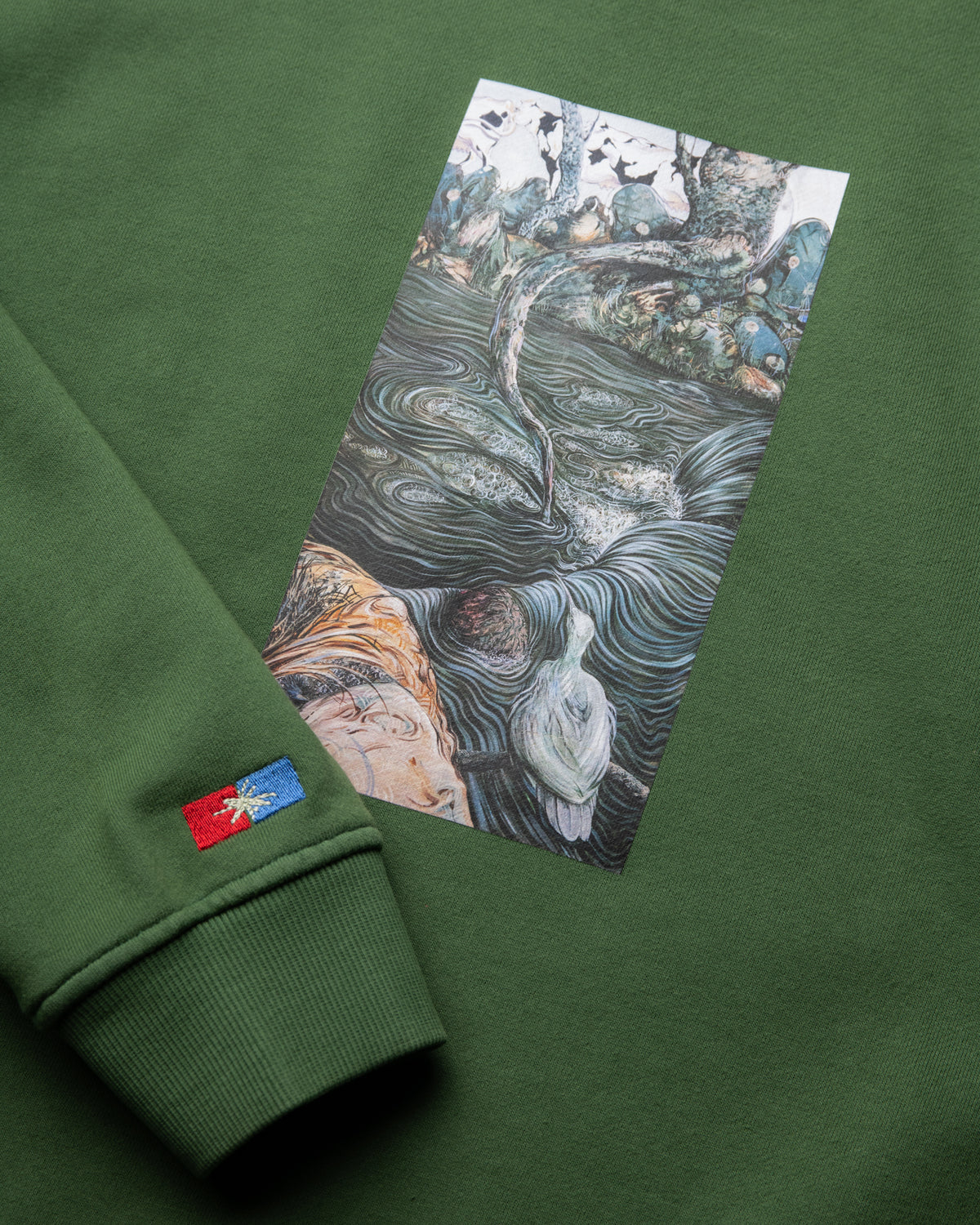 A close-up of an olive green sweatshirt featuring a rectangular graphic print of a classical painting depicting a woman in a flowing river, with a small red and blue embroidered logo on the cuff.