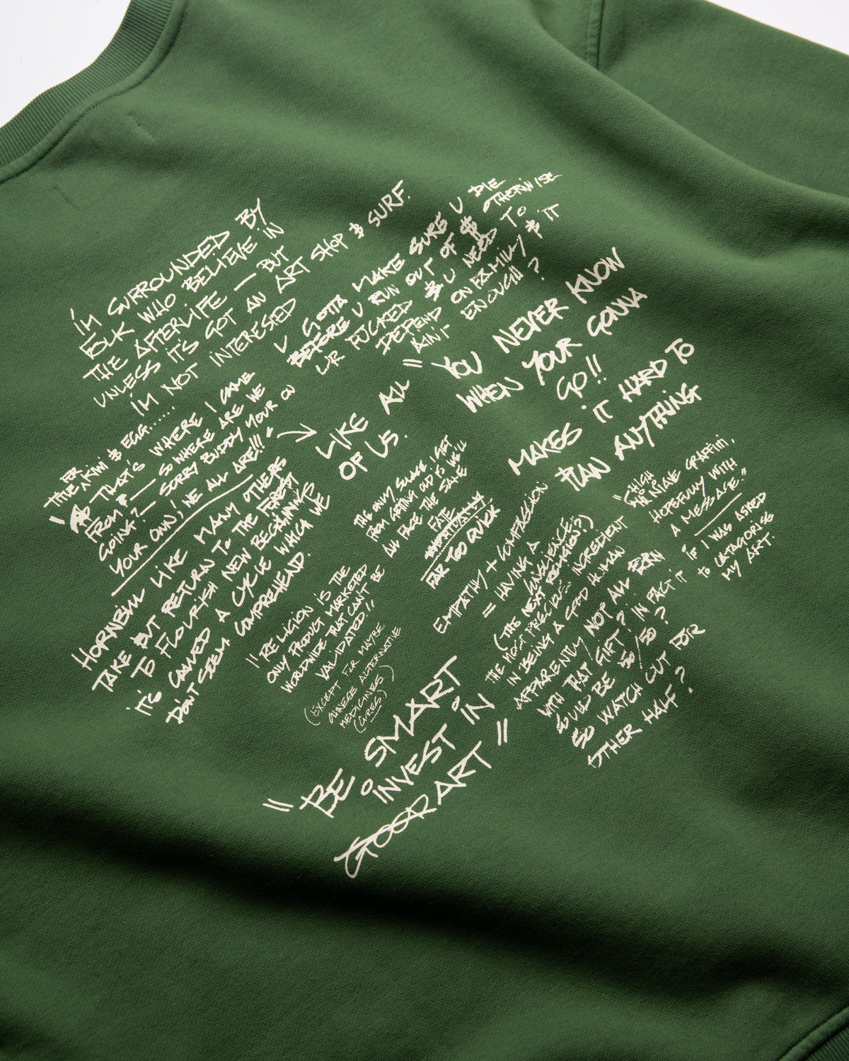 An olive green sweatshirt featuring a large white handwritten-style text collage on the front, photographed close-up against a neutral light background.