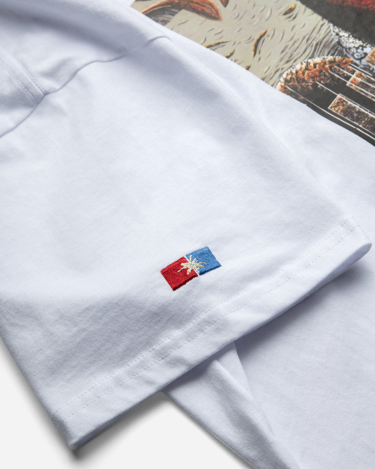 Close-up of a white short-sleeve garment, likely shorts or pajama pants, showing a small embroidered flag patch on the sleeve. The patch is red and blue with a white star-like emblem in the center.