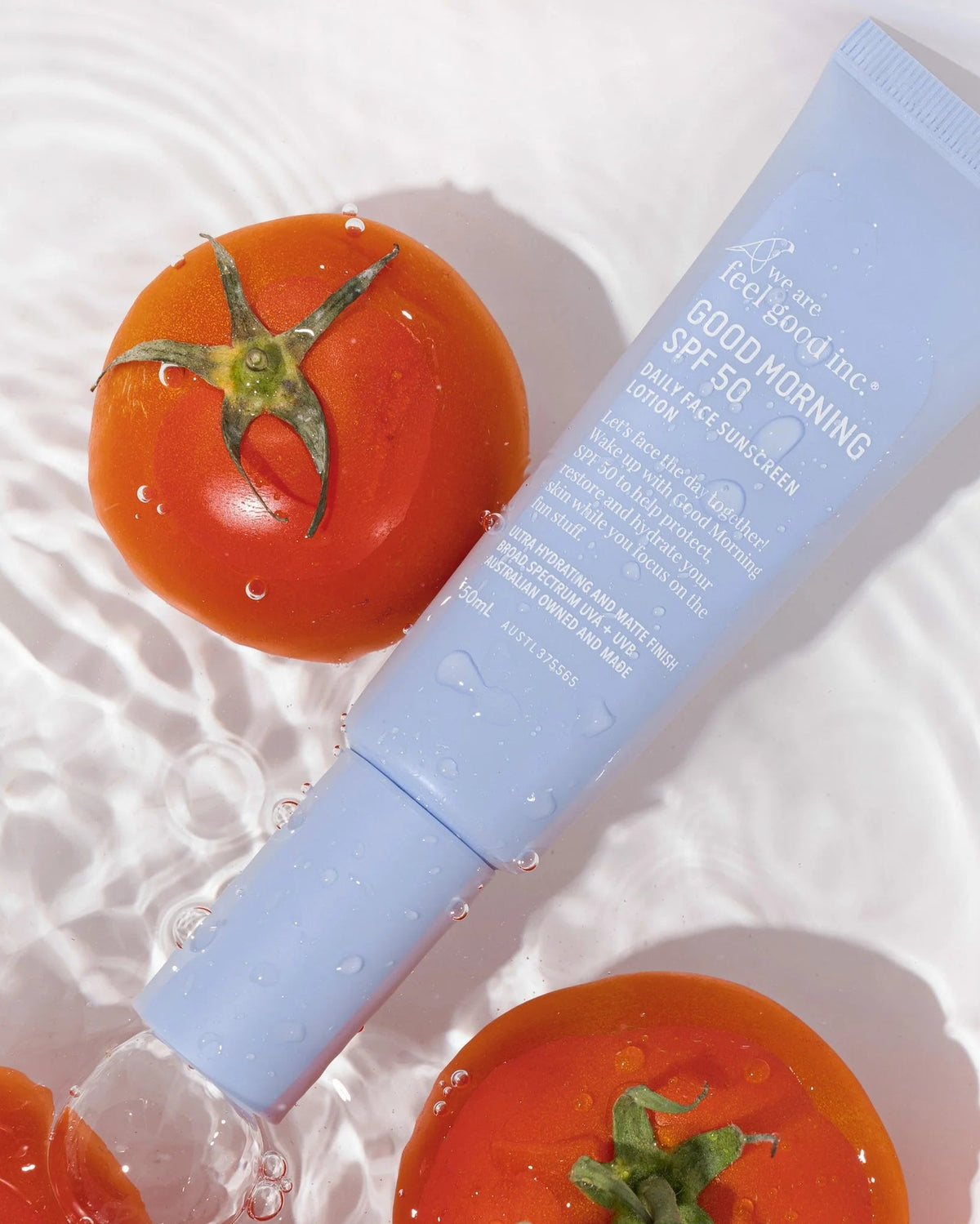 A light blue tube of 'feel free' brand Good Morning SPF 50 sunscreen lies diagonally on a white surface with ripples and water droplets, surrounded by fresh red tomatoes.