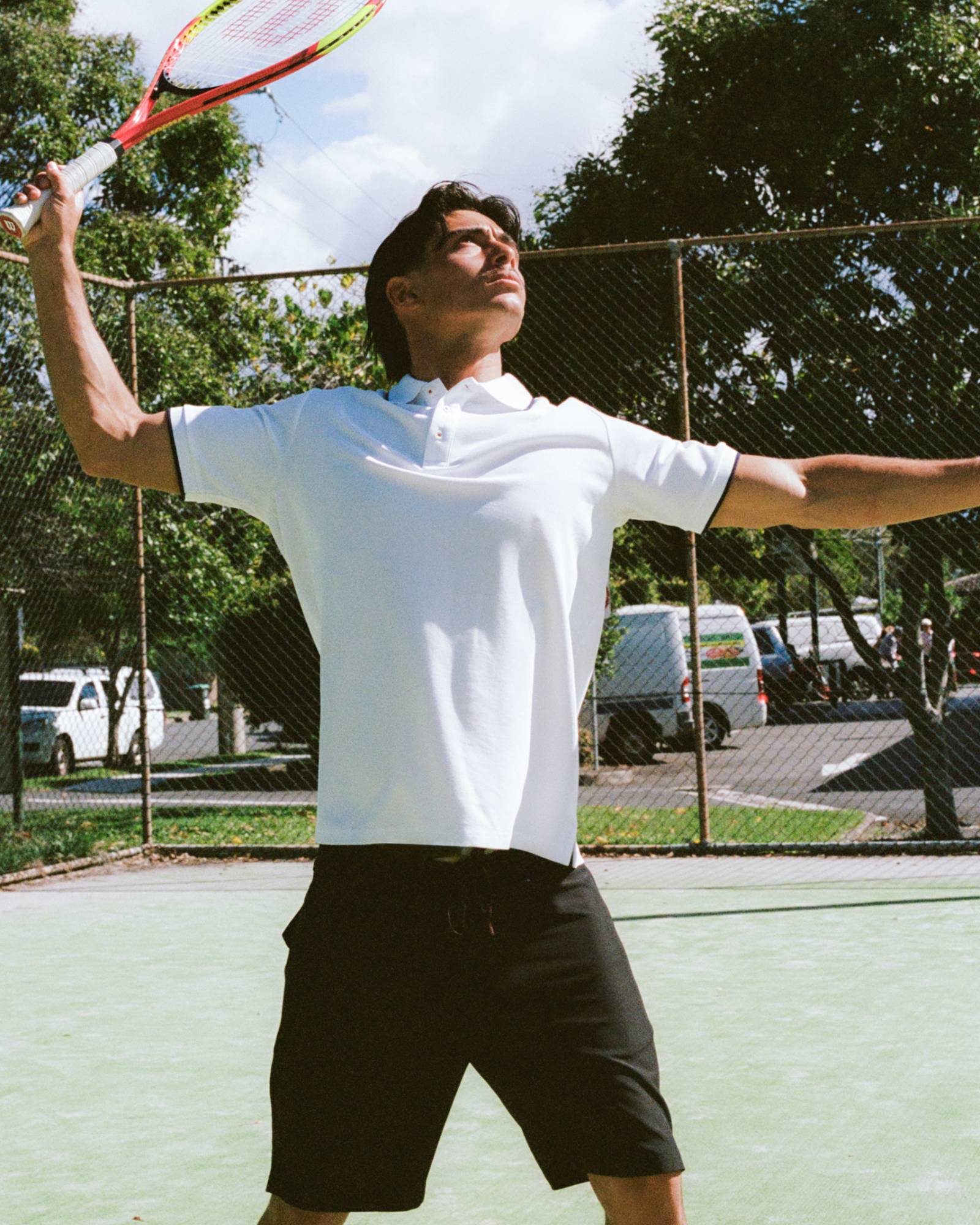Man playing tennis on a court with trees and vehicles in the background wearing a white polo