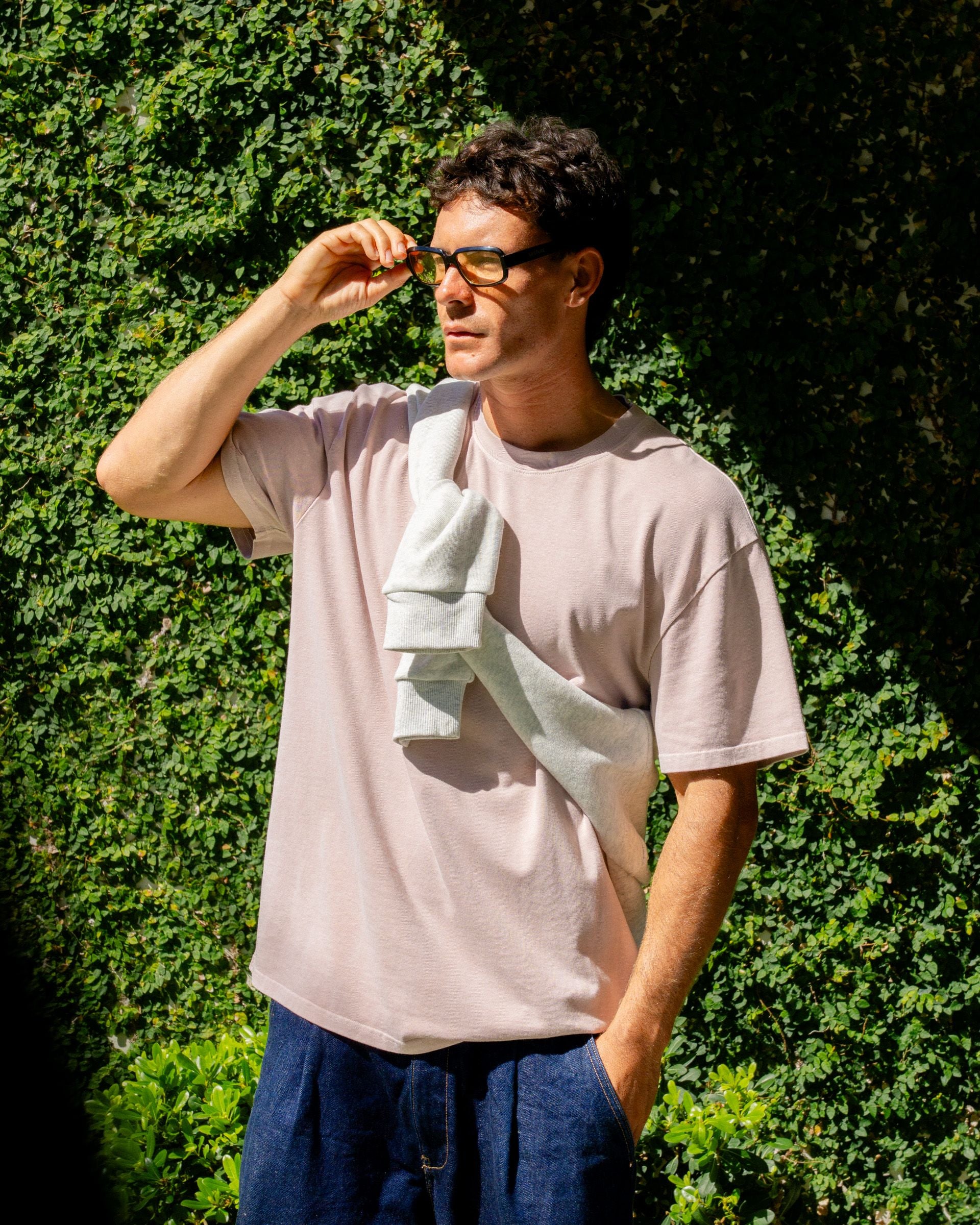 Man wearing a light pink t-shirt and blue shorts, standing outdoors with greenery in the background.