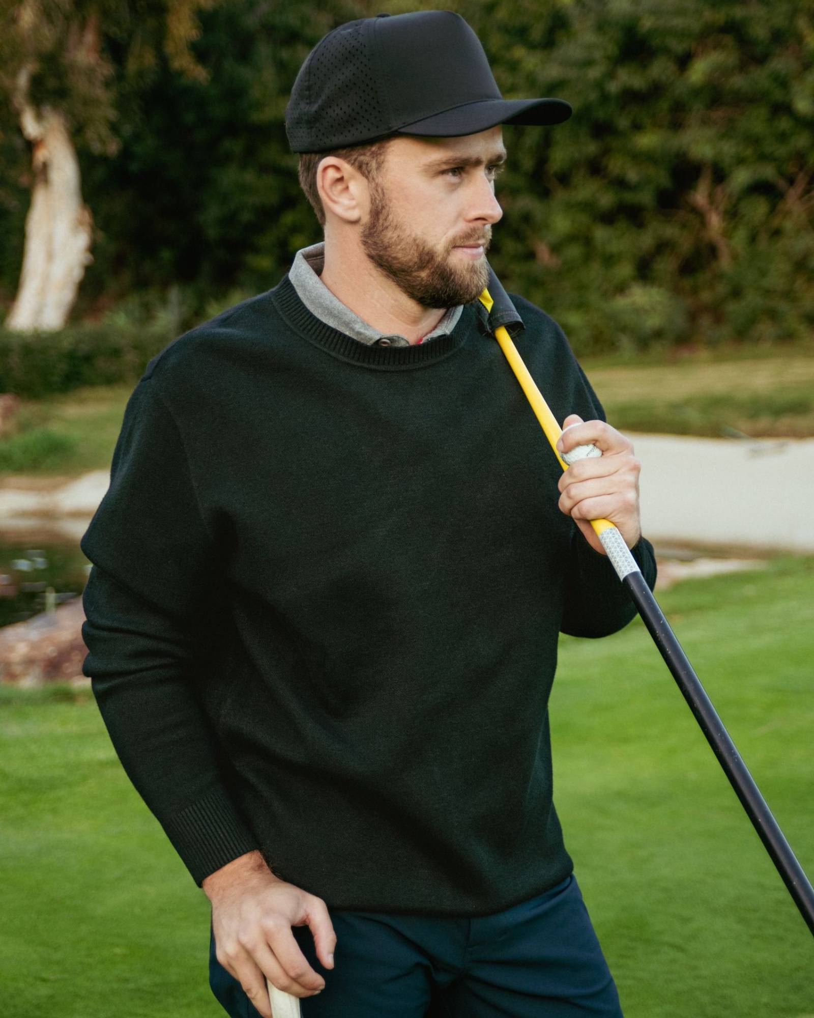 Man holding a golf club on a golf course wearing a black knit sweater