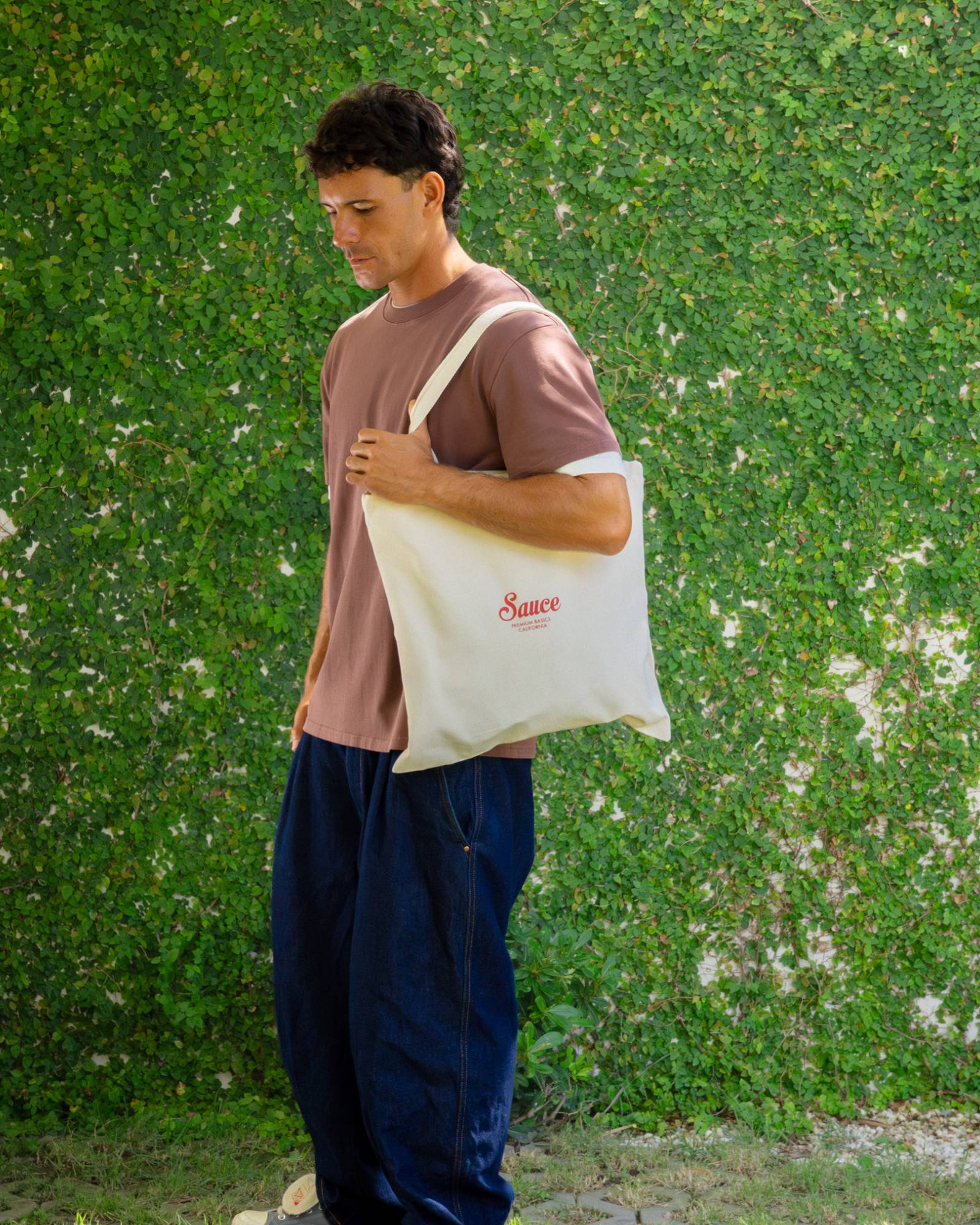 Man holding a beige tote bag with 'Sauce' branding against a green wall.