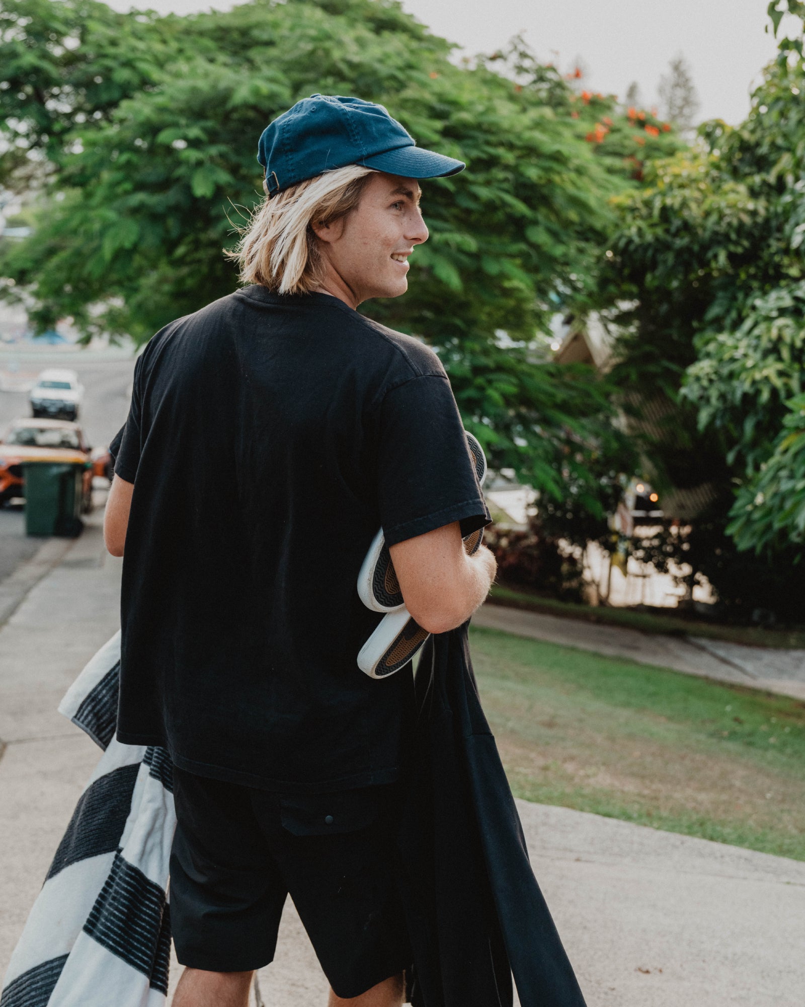 Person wearing a black t-shirt and cap, standing outdoors with trees and cars in the background