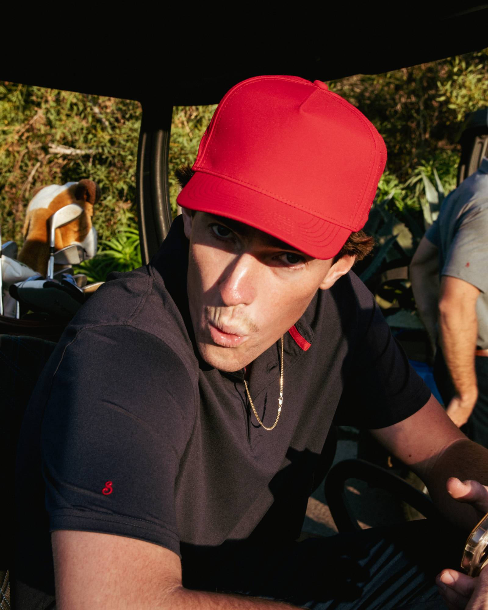 Person wearing a red cap and black shirt sitting inside a vehicle with greenery in the background
