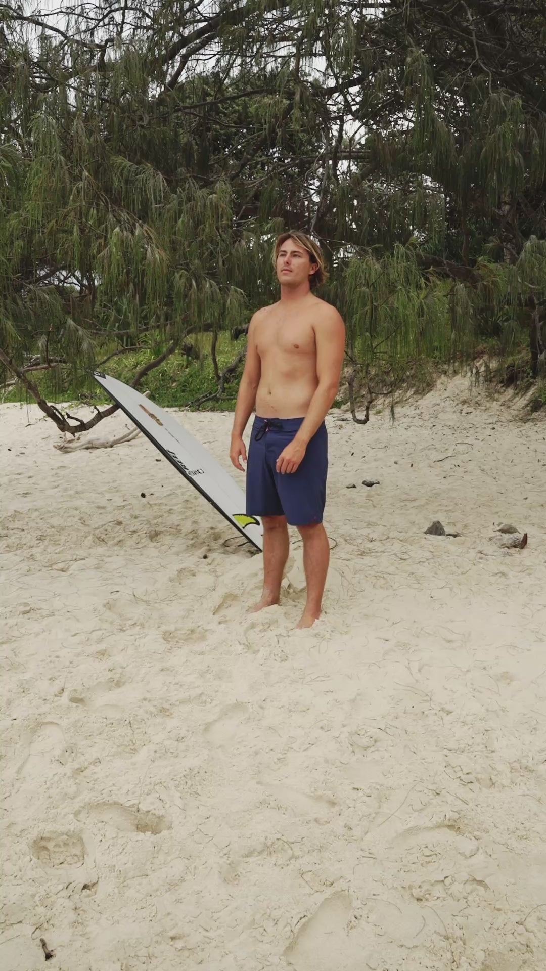 Person wearing blue boardshorts standing on sand with greenery in background