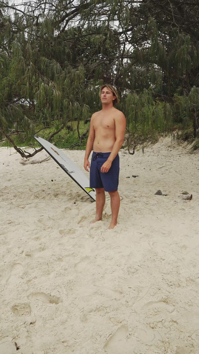 Person wearing blue boardshorts standing on sand with greenery in background