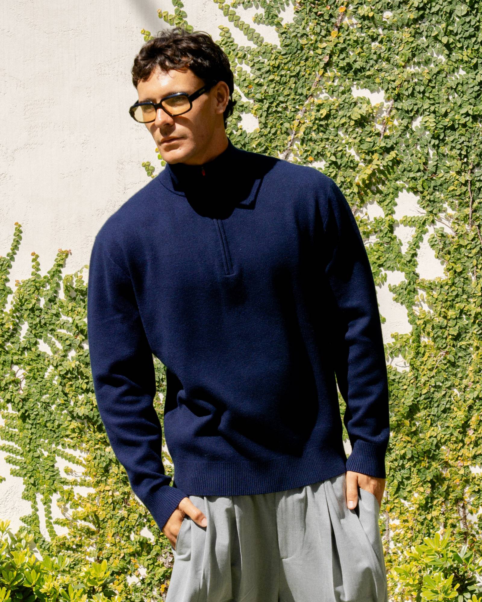 Man wearing a navy sweater and light gray pants standing against a green leafy background