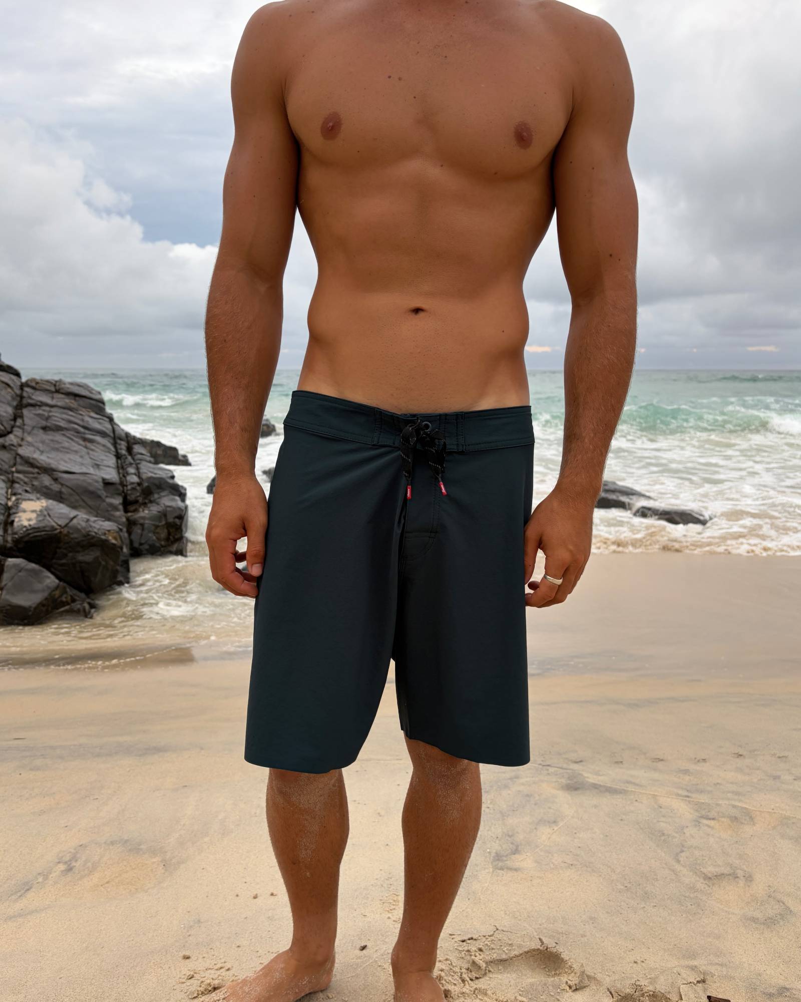 Man wearing black swim shorts standing on a beach with ocean and rocks in the background