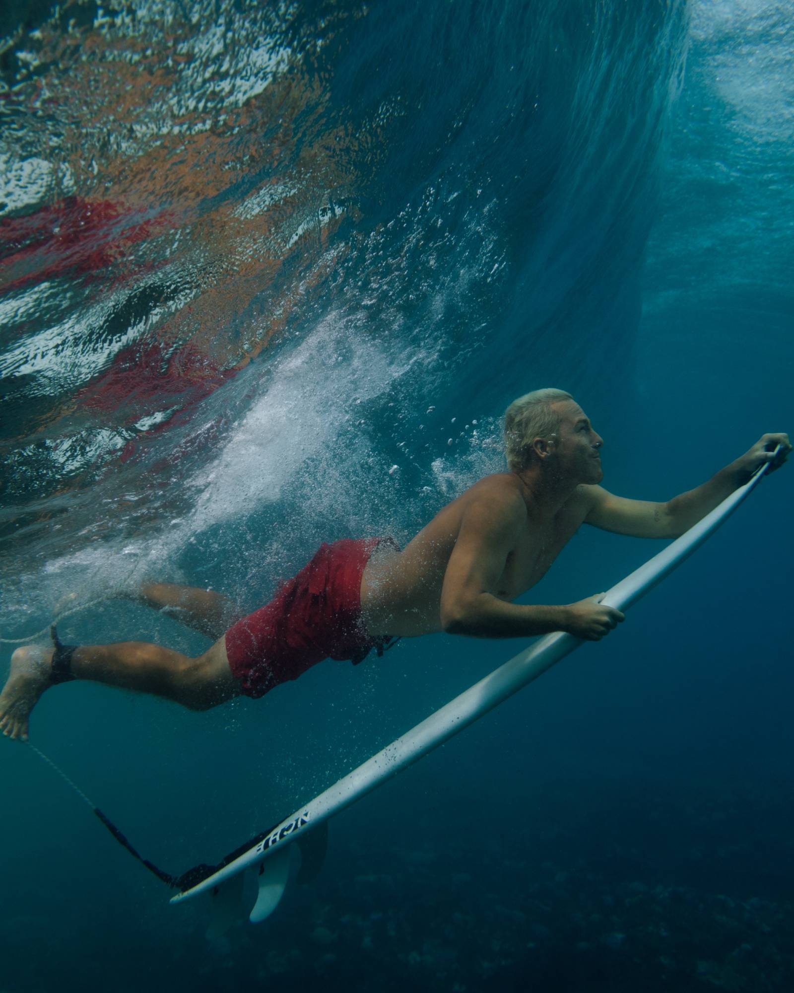 Person underwater holding a surfboard with a large wave in the background